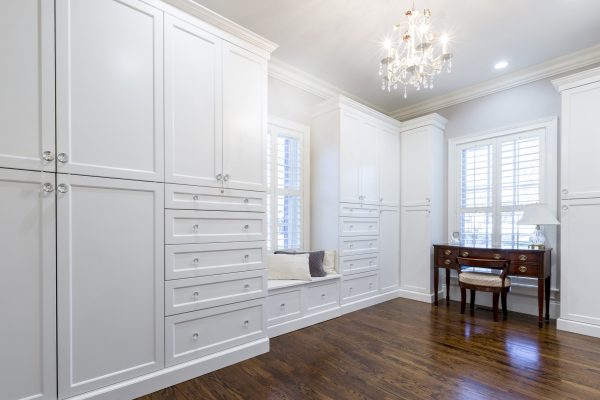 A bright, luxurious room with custom white built-in cabinetry, a window seat, dark wood floors, and a crystal chandelier.