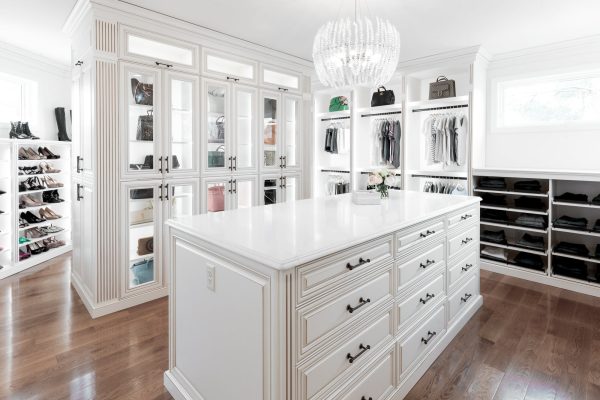 White custom closet with center island, glass-front cabinets for bags, shelves for shoes, and hanging clothes.