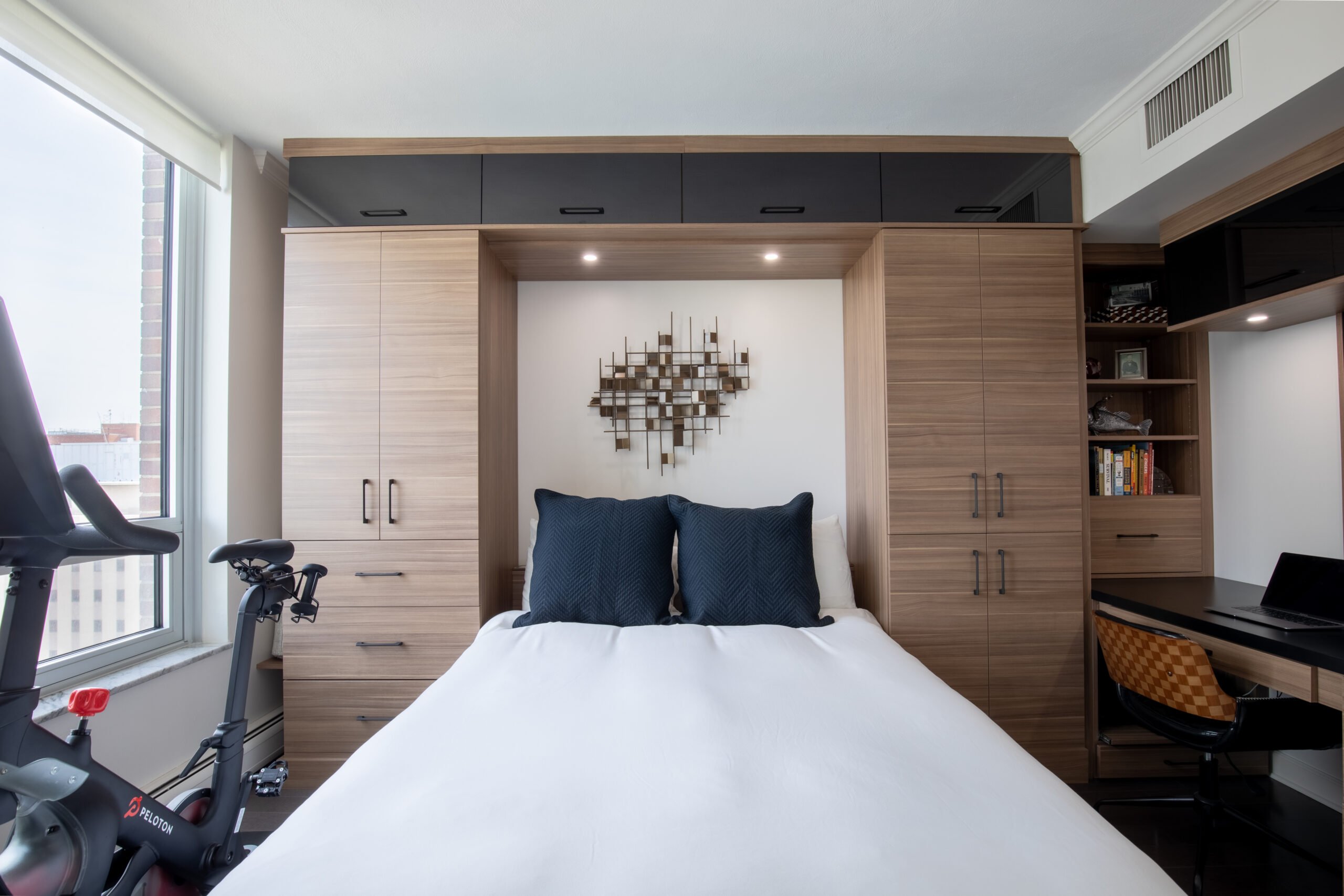 Modern custom-built bedroom with a bed bordered by warm wood cabinetry, a Peloton bike by the window and a desk.