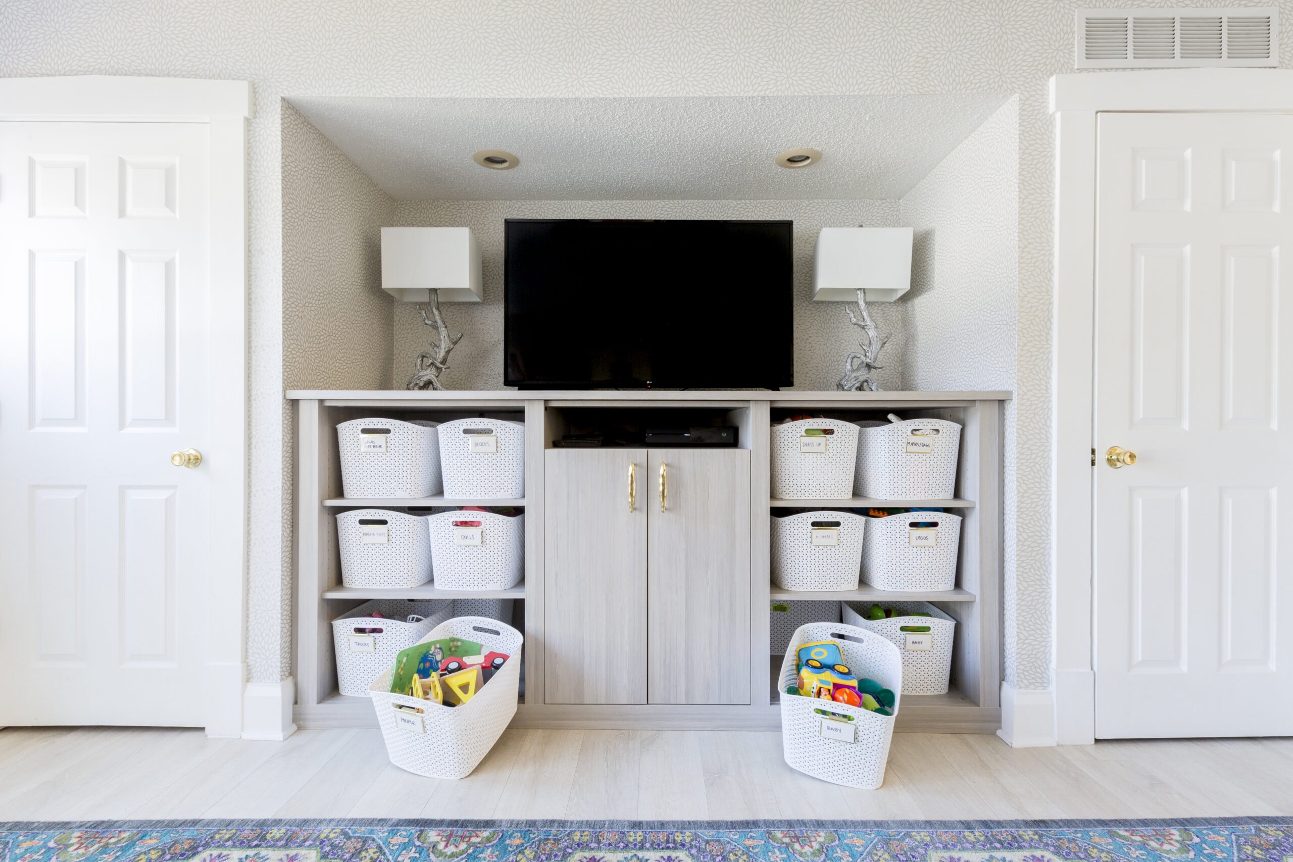 Built-in entertainment center with TV, white patterned wallpaper in a playroom, toy storage baskets, and light wood floors.