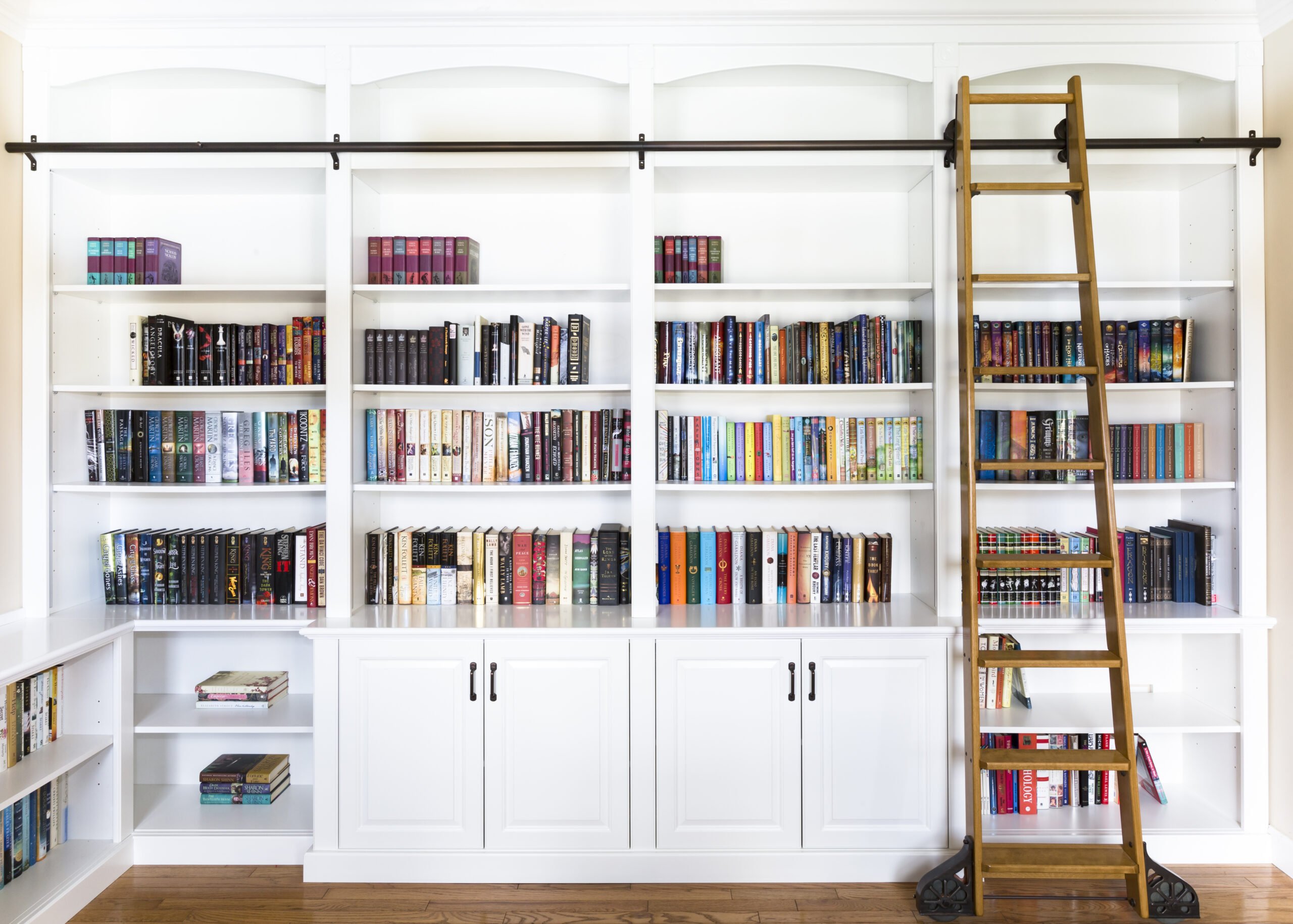 White built-in bookshelves with a wooden rolling ladder on a dark rod, filled with colorful books.