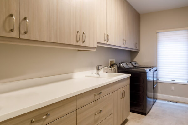 Modern laundry room with light wood cabinets, white countertop, sink, black washer and dryer, and a window with blinds.