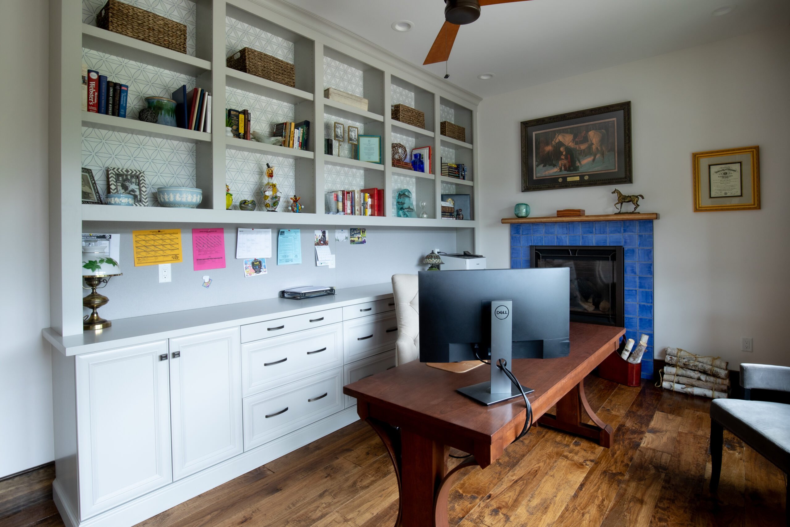 A cozy home office featuring built-in gray bookshelves with geometric patterned backs, a blue-tiled fireplace, and a wooden desk.