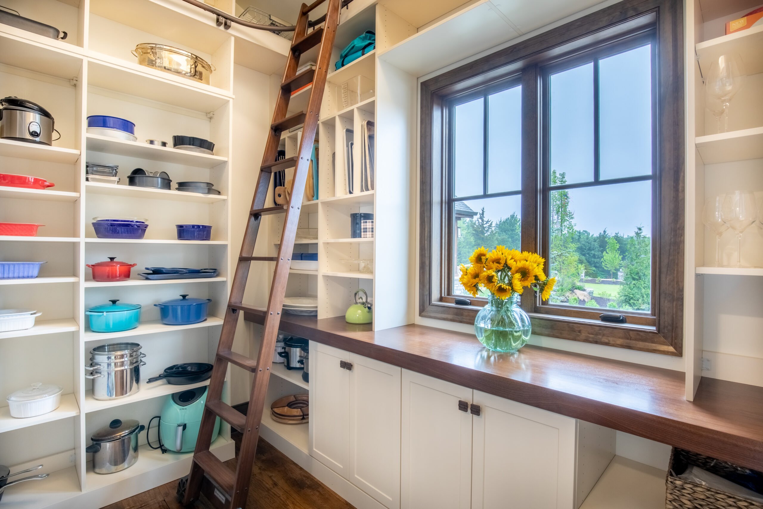 A cozy pantry with white shelves full of kitchenware, a rolling library ladder, and a window with sunflowers overlooking a garden.
