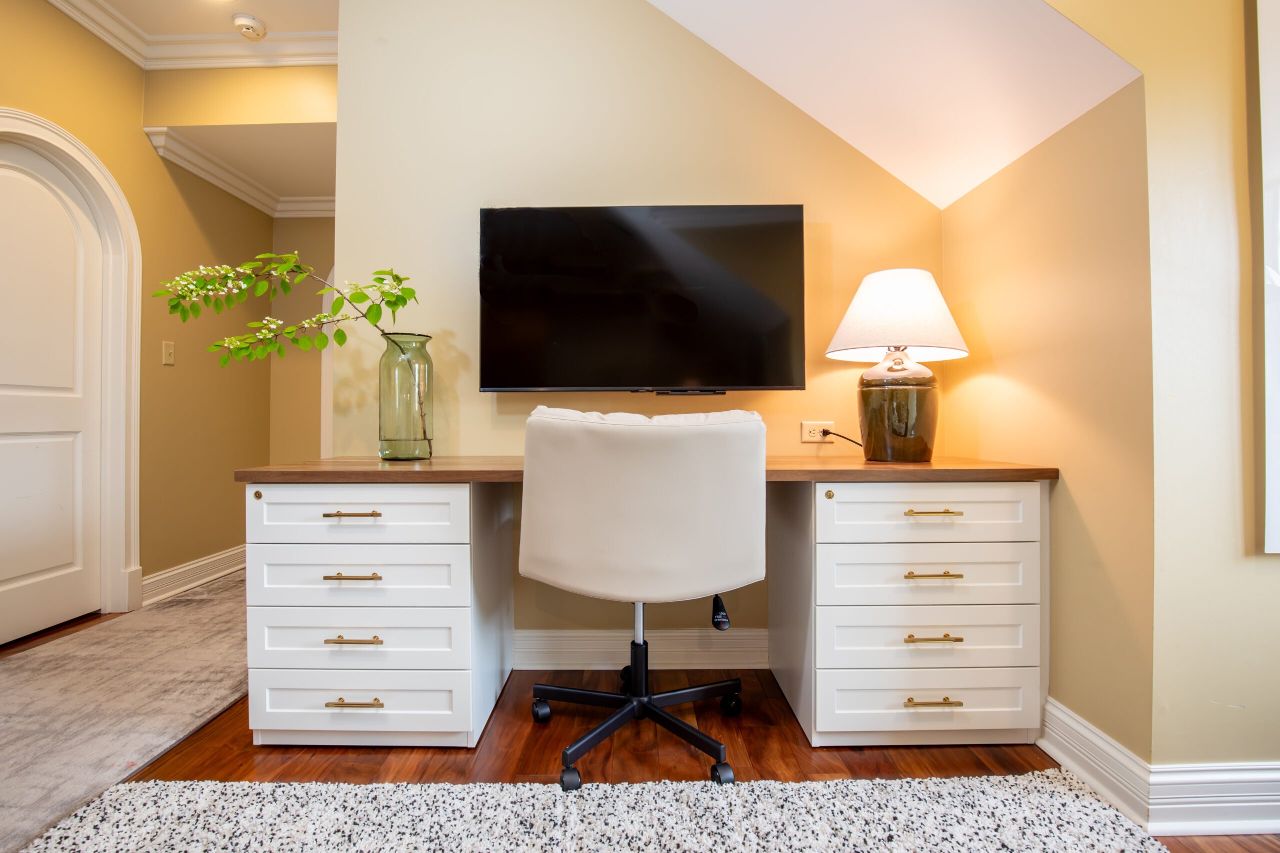 A cozy home office nook with a wooden desk, white drawer cabinets, a white chair, and a mounted TV.