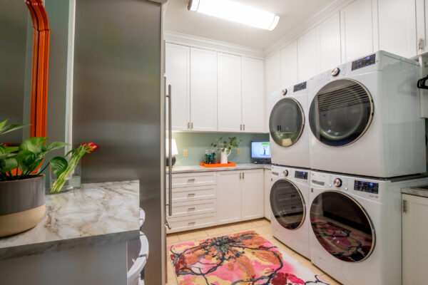 Bright, modern laundry room with white cabinets, light blue wall, stacked washer/dryer, marble countertops, and a vibrant rug.