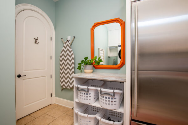 Laundry room with light green walls, a white arched door, and a zigzag ironing board. An orange mirror is above white baskets and a steel appliance.