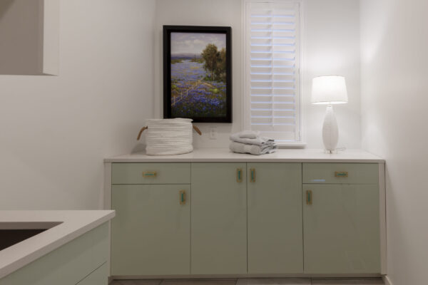 Laundry room with light green cabinets, white countertops, bluebonnet painting, white hamper, folded towels, and a decorative white lamp.