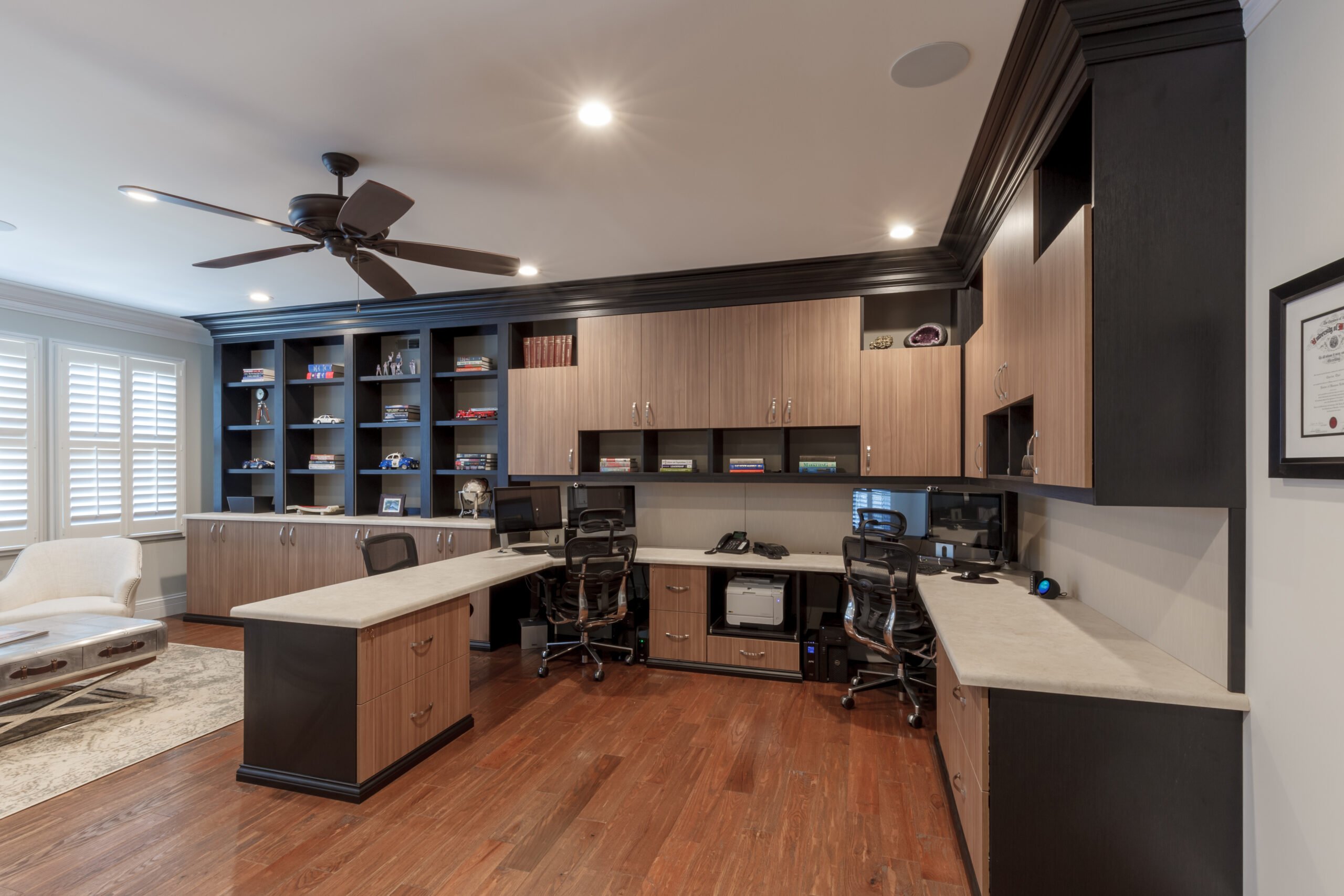 Home office with black and wood custom cabinetry, neutral countertops, dark wood flooring, and a ceiling fan.