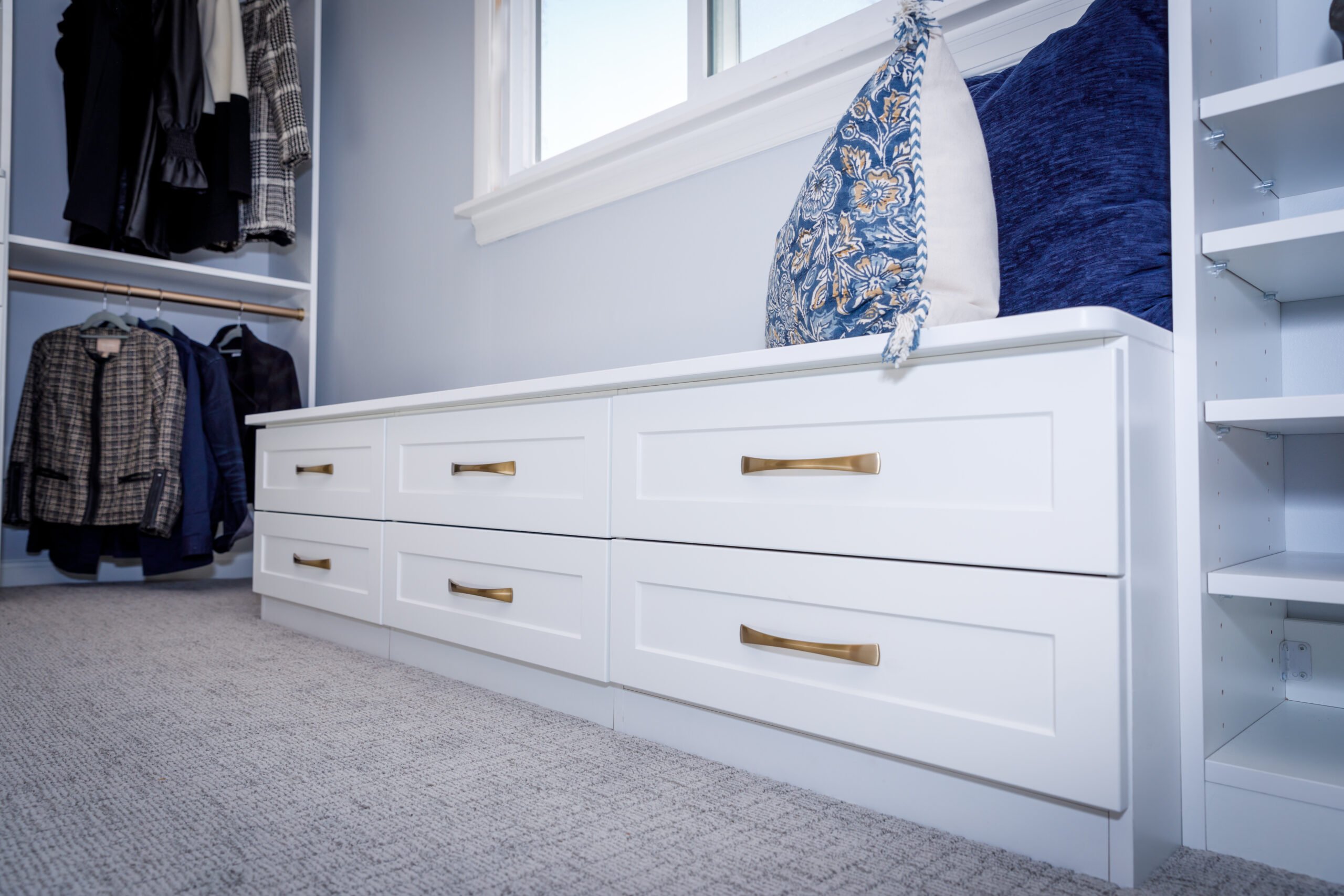 White built-in storage bench with gold handles, blue cushions, next to a window and hanging clothing in a closet.