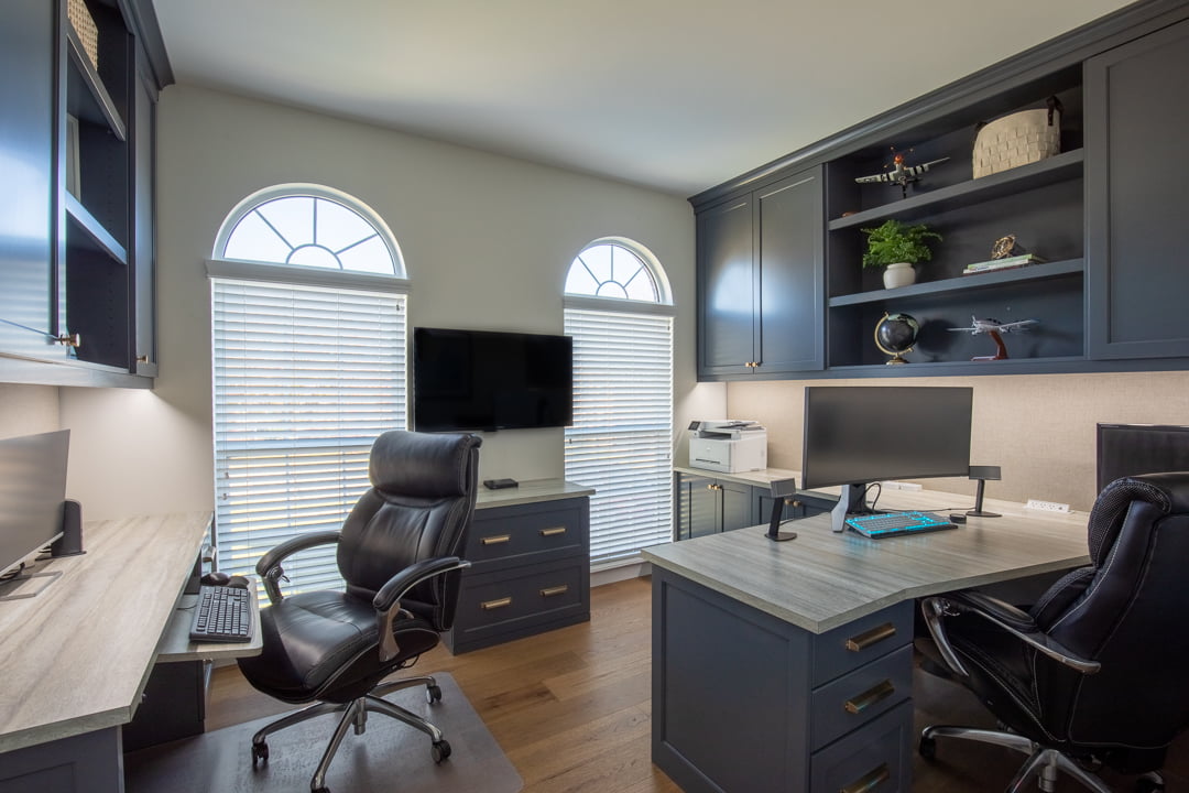 A stylish home office with dark blue cabinets, light wood counters, two black office chairs, arched windows, and decorative shelves.