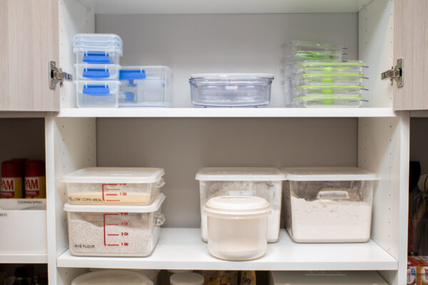 Organized kitchen cabinet with clear food storage containers. Top shelf has empty containers, bottom shelf holds yellow corn meal and rye flour.