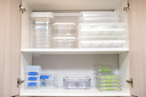 Open kitchen cabinet with clear plastic food storage containers. Some have white, blue, or green lids.