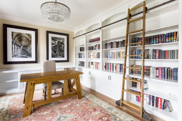 Luxurious home office with white built-in bookshelves, rolling ladder, wooden desk, and ornate chandelier.