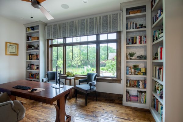 Spacious home office with hardwood floors, dark wood desk, built-in bookshelves, and large window overlooking trees.