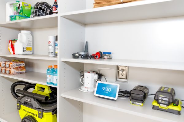 White shelves in a garage. Top left shelf: cleaning supplies, hockey helmet. Next shelf: cleaner, spray paint, drinks. Bottom left: shop vacuum. Bottom right: smart display, charging batteries.