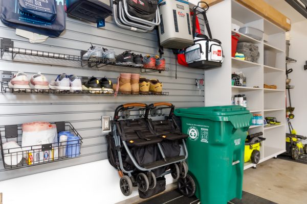 Organized garage with shoes on wall-mounted shelves, a double stroller, green trash bin, and white storage shelves.