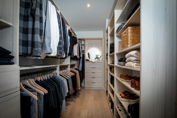 Light wood walk-in closet with hanging clothes, shelves, folded items, and a dresser with a round mirror.