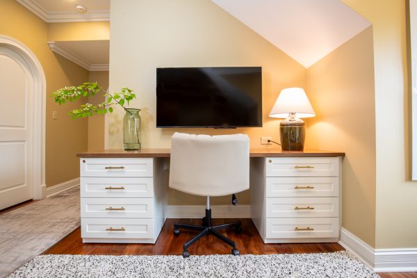 Home office with wooden desk, white drawers, TV, light beige chair, and lamp.