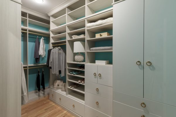 Full walk-in closet with beige cabinetry & turquoise walls. Gold hanging rods store shirts & pants. Shelves hold shoes, bags, & towels.