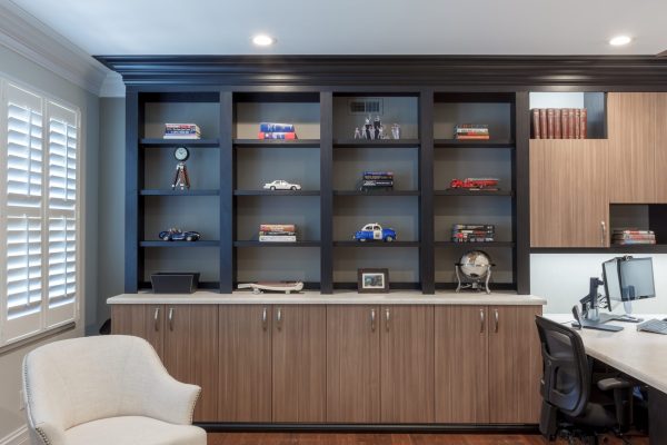 Home office with dark built-in shelves displaying books and collectibles, cream cabinetry, and a light desk with dual monitors.