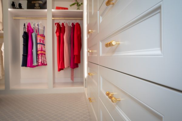 White custom closet drawers with gold hardware in the foreground, with colorful dresses hanging in the background.