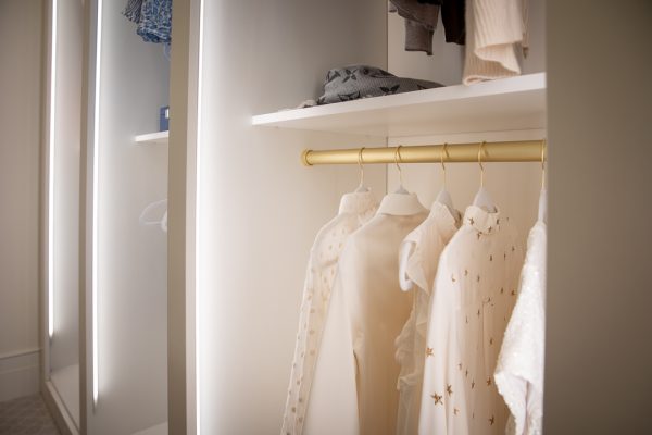White closet with golden clothes rail, housing hanging garments and folded clothes on a shelf above.