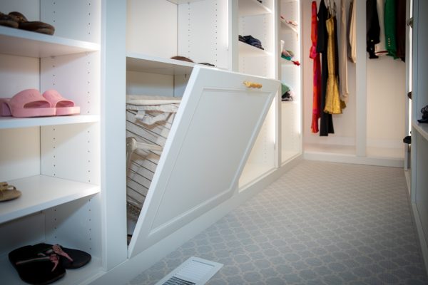 White custom closet with built-in tilt-out laundry hamper and hanging clothes, light blue carpet.