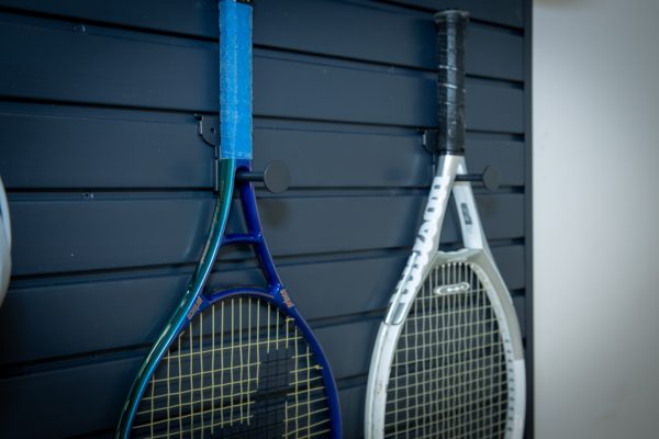 Two tennis rackets, blue and silver, hanging on a dark slat wall with hooks.