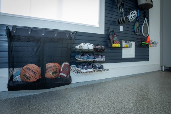 Garage storage with black slatwall, mesh sports bag with basketballs, football, tennis shoes on wire racks, and tennis rackets.