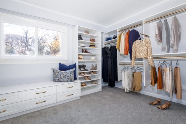 Spacious walk-in closet with white shelving, hanging clothes, shoe racks, bench seating, window, and leopard heels on carpet.