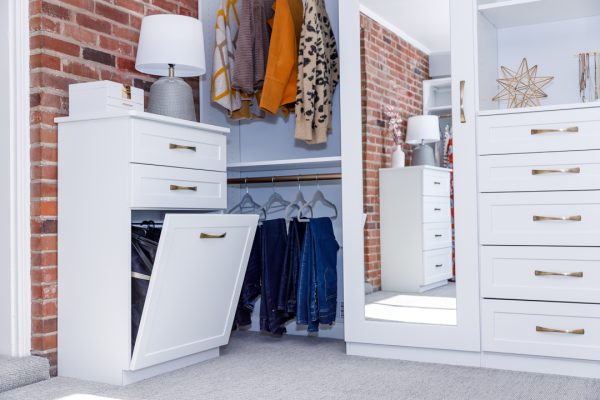 White closet system with pull-out hamper, drawers, hanging clothes, and mirror by a brick wall.