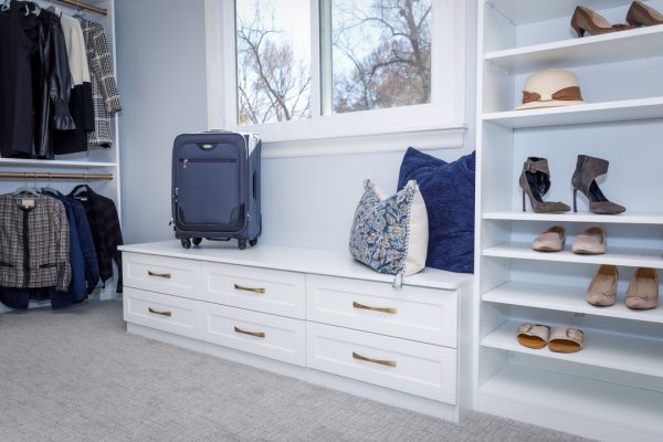A custom walk-in closet with white shelving, hanging clothes, drawers, shoe racks, a window, and a small blue suitcase.