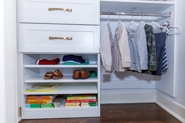 White custom closet with drawers, open shelves of folded clothes, shoes, and children's books, plus a hanging rod of clothes.