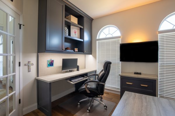 Home office with custom gray cabinets, built-in desk, black monitor, chair, and arched windows.