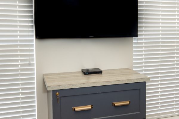 Wall-mounted TV above a gray wooden chest with two drawers and brass handles, light-colored top.
