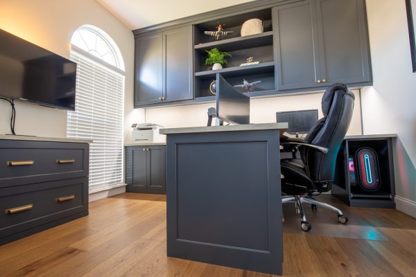 Home office with dark gray cabinets, hardwood floors, a TV, arched window, and a gaming computer with LED lights.
